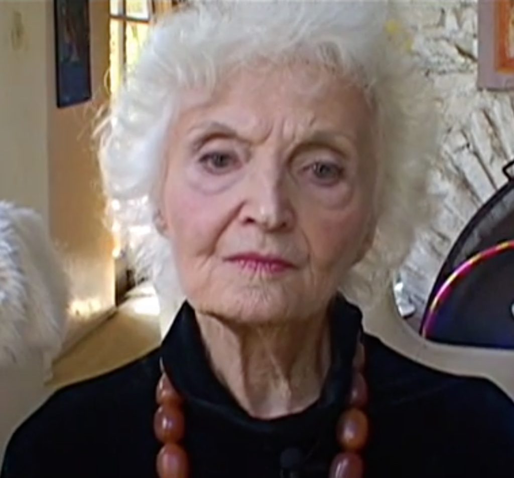 Elder Laura Huxley looking at the camera wearing a signature chunky necklace with a hula hoop behind her leaning against a fireplace.